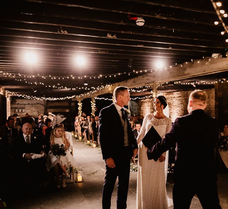 Bride in Lenora Dress by Wtoo Watters with Lace Sleeves and Keyhole Back | Prudence Halo Headpiece by Kelly Spence Wed | Floor Length Veil | Groom in Navy Suit with Grey Waistcoat from Jack Bunneys | Fairy Light Décor | Candle and Foliage Lined Aisle | Exposed Brick Industrial Venue | The Electrician’s Shop at Trinity Buoy Wharf | String Lights, Perspex Table Signs and Paper Cranes for Industrial Wedding | Frankee Victoria Photography