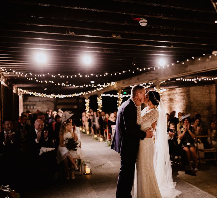 Bride in Lenora Dress by Wtoo Watters with Lace Sleeves and Keyhole Back | Prudence Halo Headpiece by Kelly Spence Wed | Floor Length Veil | Groom in Navy Suit with Grey Waistcoat from Jack Bunneys | Fairy Light Décor | Candle and Foliage Lined Aisle | Exposed Brick Industrial Venue | The Electrician’s Shop at Trinity Buoy Wharf | String Lights, Perspex Table Signs and Paper Cranes for Industrial Wedding | Frankee Victoria Photography