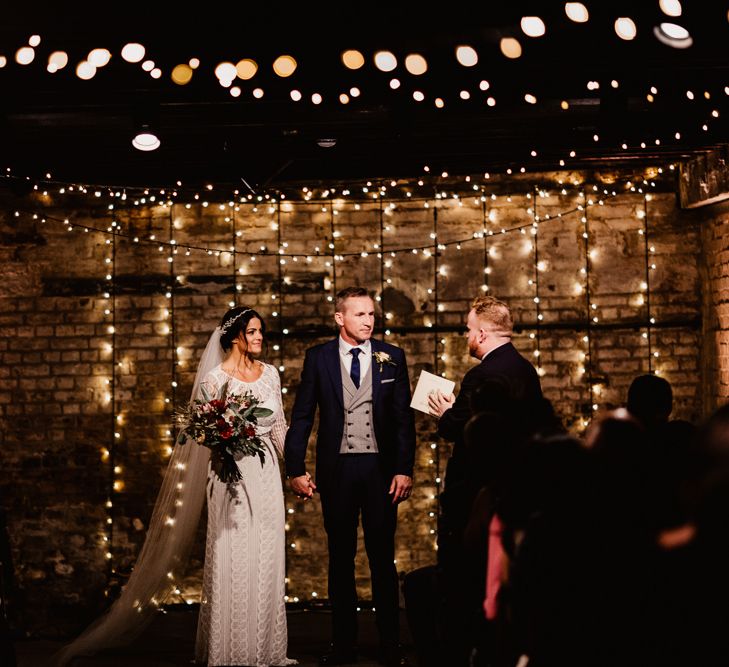 Bride in Lenora Dress by Wtoo Watters with Lace Sleeves and Keyhole Back | Prudence Halo Headpiece by Kelly Spence Wed | Floor Length Veil | Burgundy, Blush and White Flowers with Foliage and Ferns | Groom in Navy Suit with Grey Waistcoat from Jack Bunneys | Fairy Light Canopy | Exposed Brick Industrial Venue | The Electrician’s Shop at Trinity Buoy Wharf | String Lights, Perspex Table Signs and Paper Cranes for Industrial Wedding | Frankee Victoria Photography