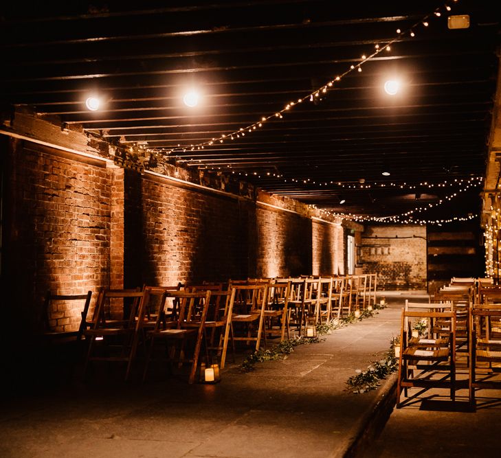 Fairy Light Décor | Candle and Foliage Lined Aisle | Exposed Brick Industrial Venue | The Electrician’s Shop at Trinity Buoy Wharf | String Lights, Perspex Table Signs and Paper Cranes for Industrial Wedding | Frankee Victoria Photography