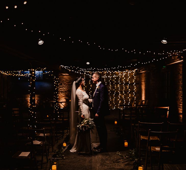 Bride in Lenora Dress by Wtoo Watters with Lace Sleeves and Keyhole Back | Prudence Halo Headpiece by Kelly Spence Wed | Floor Length Veil | Burgundy, Blush and White Flowers with Foliage and Ferns | Groom in Navy Suit with Grey Waistcoat from Jack Bunneys | Fairy Light Canopy | Candle and Foliage Lined Aisle | Exposed Brick Industrial Venue | The Electrician’s Shop at Trinity Buoy Wharf | String Lights, Perspex Table Signs and Paper Cranes for Industrial Wedding | Frankee Victoria Photography