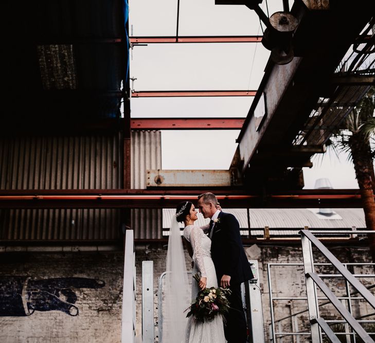 Bride in Lenora Dress by Wtoo Watters with Lace Sleeves and Keyhole Back | Prudence Halo Headpiece by Kelly Spence Wed | Floor Length Veil | Burgundy, Blush and White Flowers with Foliage and Ferns | Groom in Navy Suit with Grey Waistcoat from Jack Bunneys | String Lights, Perspex Table Signs and Paper Cranes for Industrial Wedding | Frankee Victoria Photography