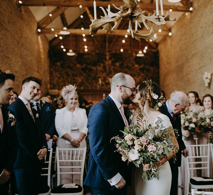 Bride in Pronovias Dralan Wedding Dress  and Groom in Navy Blue Moss Bros. Suit Kissing at the Altar