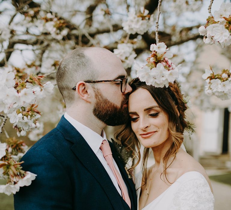 Bride in Pronovias Dralan Wedding Dress and Groom in Navy Moss Bros. Suit with Pink Tie Standing in Front of a Blossom Tree