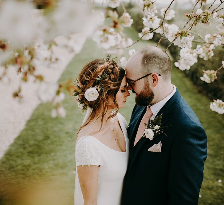 Bride in Pronovias Dralan Wedding Dress and Groom in Navy Moss Bros. Suit Cuddling in Front of a Blossom Tree