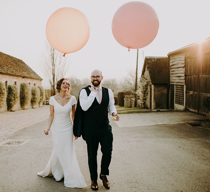 Bride in Pronovias Dralan Wedding Dress and Groom in Navy Moss Bros. Suit Holding Giant Pink and Peach Balloons