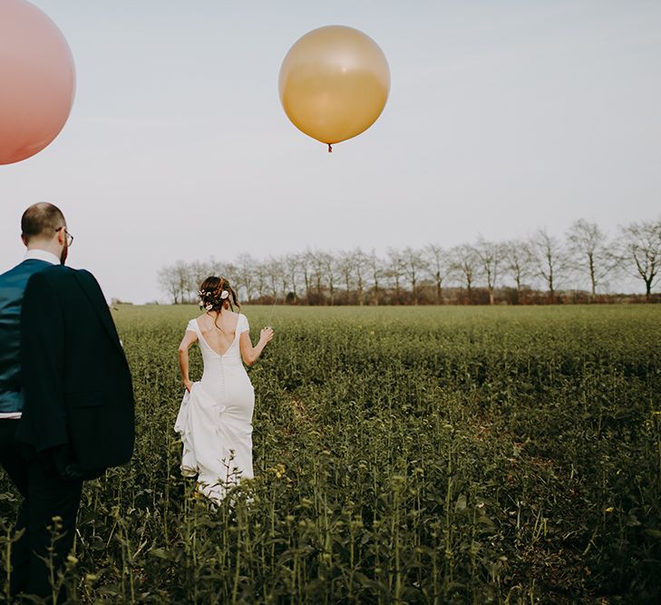 Bride in Pronovias Dralan Wedding Dress and Groom in Navy Moss Bros. Suit Walking Through a Field Holding Giant Pink and Peach Balloons