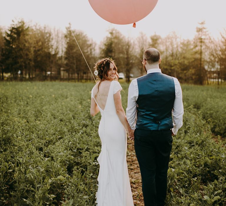 Bride in Pronovias Dralan Wedding Dress and Groom in Navy Moss Bros. Suit Walking Through a Field Holding Giant Balloons