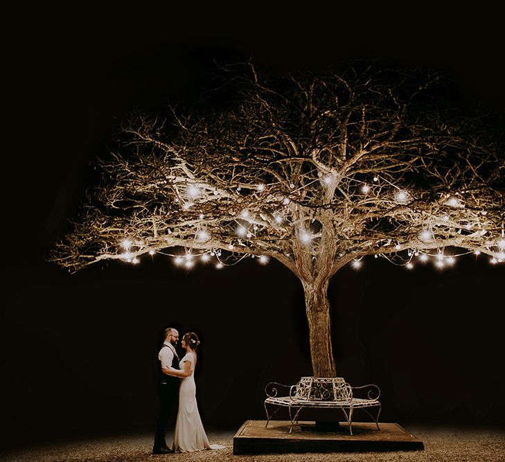 Bride in Pronovias Dralan Wedding Dress and Groom in Moss Bros. Suit Standing by a Fairy Light Covered Tree