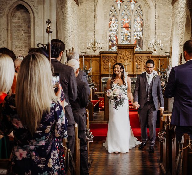 Bride wearing laced dress and pastel floral bouquet with groom wearing grey three piece suit and tie