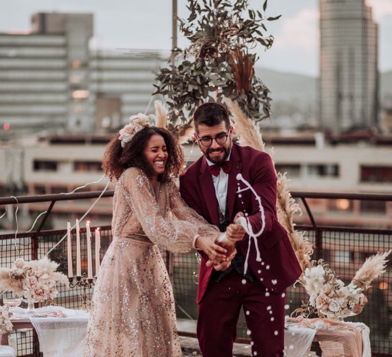 Bride in sparkle wedding dress and groom in burgundy suit popping champagne