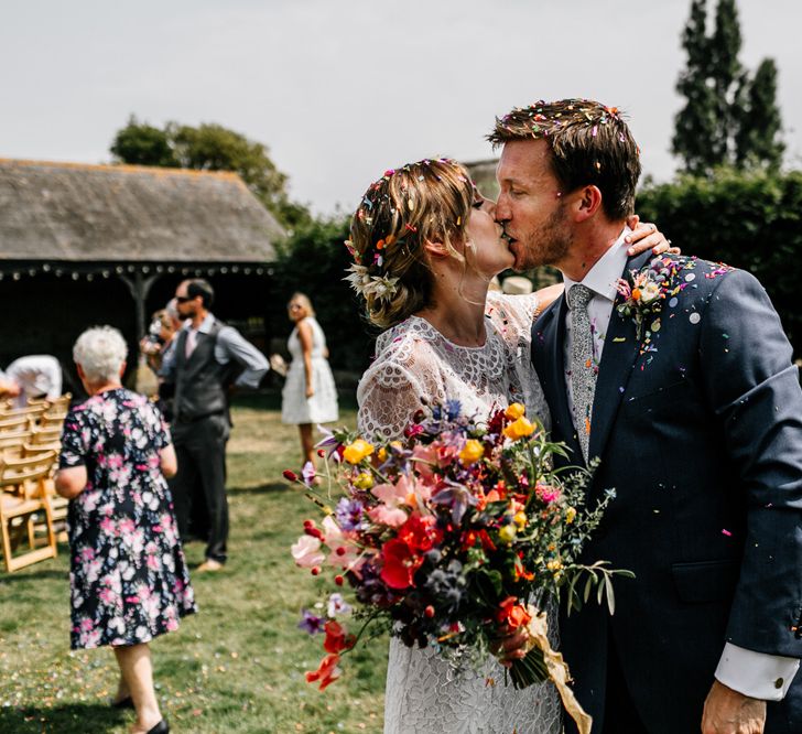 Bride in Lace Made with Love Wedding Dress and Groom in Navy Suit Kissing Covered in Confetti