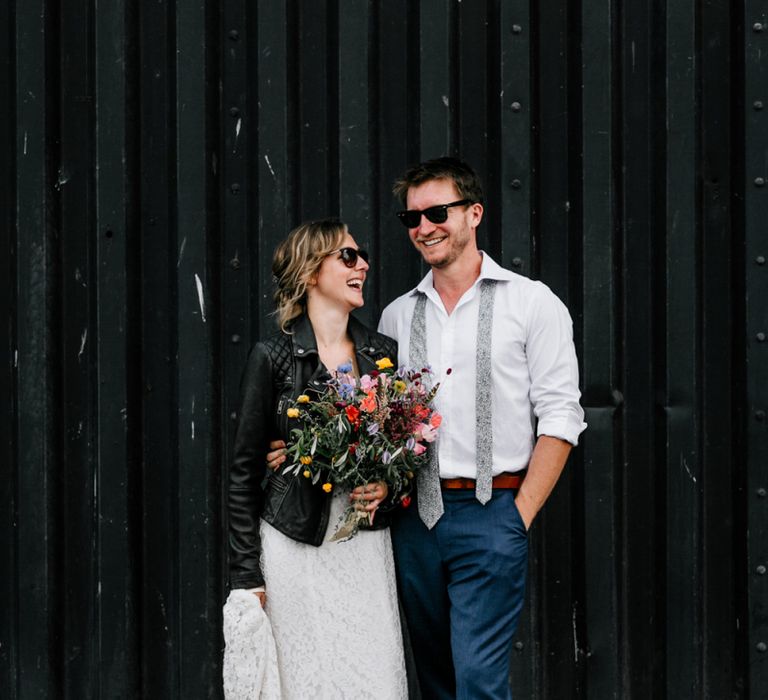 Cool Bride in Lace Made with Love Wedding Dress and Leather Jacket and Groom in Navy Trousers, Floral Tie  and Shades