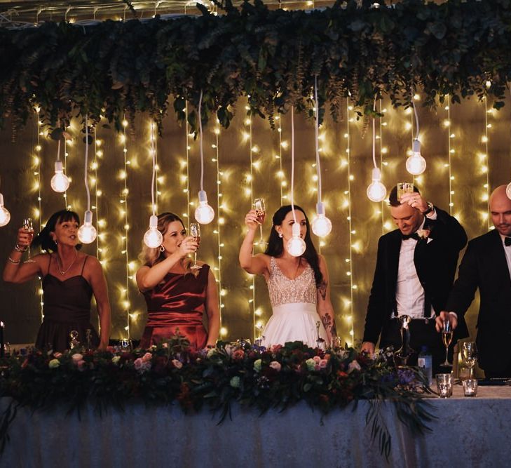 Happy Couple And Guests Toast During Wedding Speeches In Front Of Fairy Lights