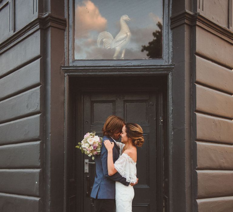 Bride in Off The Shoulder Grace Loves Lace Wedding Dress and Groom in Blue Blazer Cuddling