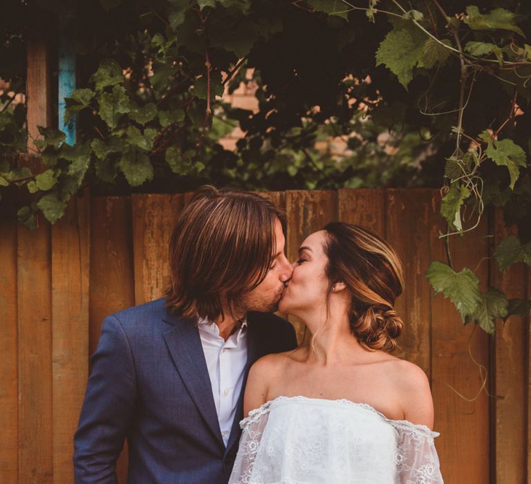 Bride in Off The Shoulder Grace Loves Lace Wedding Dress and Groom in Blue Blazer Kissing