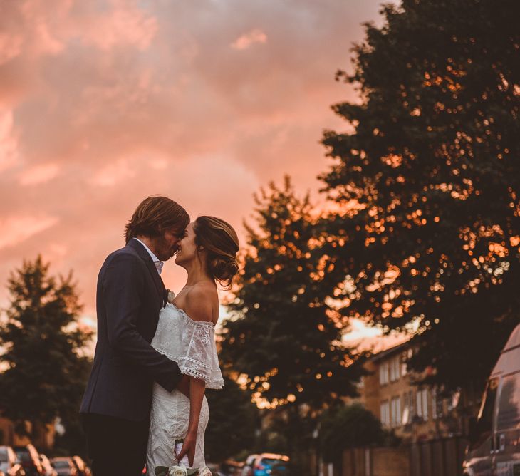 Bride in Off The Shoulder Grace Loves Lace Dress and Groom in Blue Blazer Kissing During Golden Hour