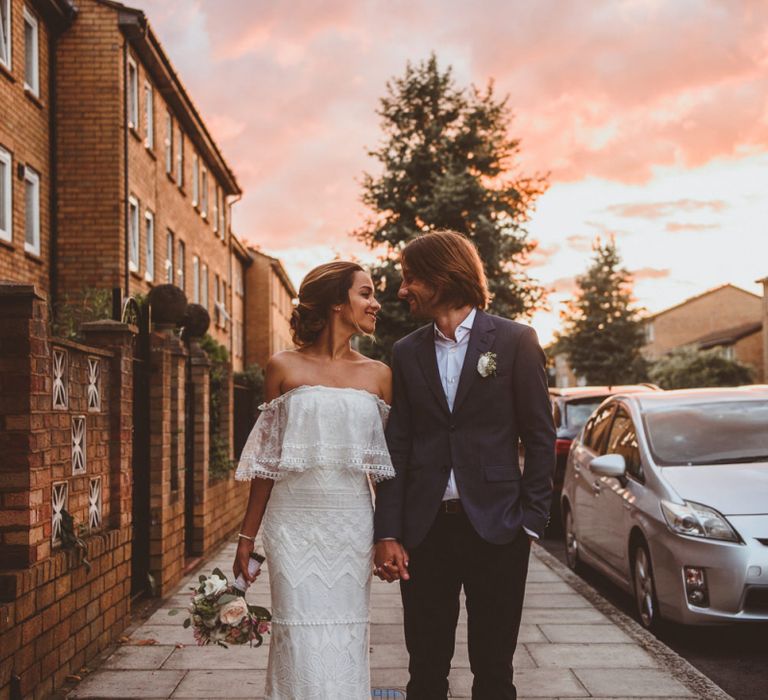 Bride in Off The Shoulder Grace Loves Lace Wedding Dress and Groom in Blue Blazer  Walking During Pink Sunset