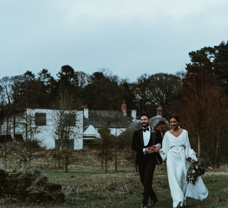 Bride and groom steal a moment at winter wedding wearing elegant Charlie Brear dress with white foliage bouquet