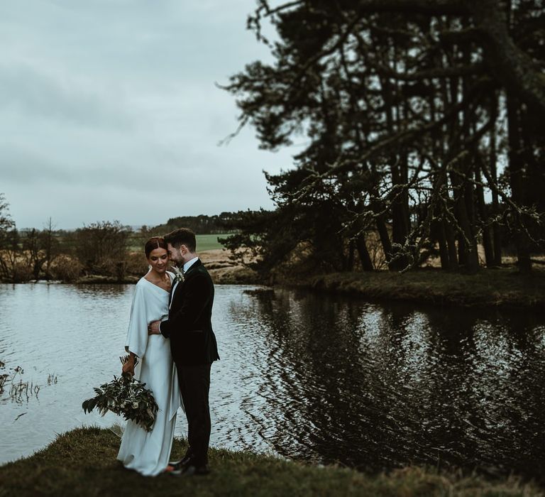 Bride and groom steal a moment at winter wedding wearing elegant Charlie Brear dress with white foliage bouquet