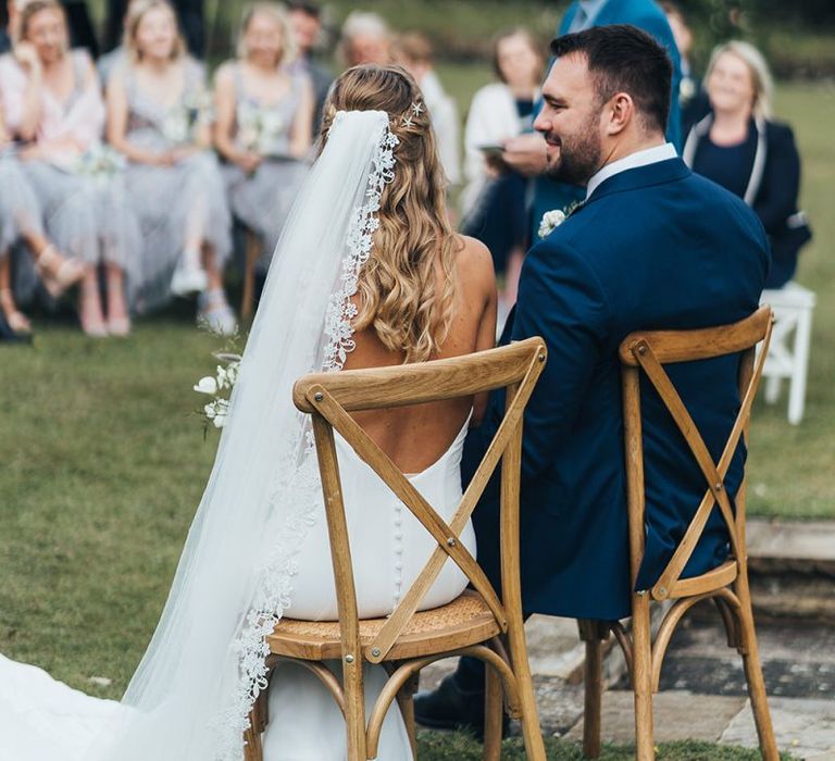 Bride in Backless Made With Love Wedding Dress and Lace Edged Veil and Groom in Navy Suit Sitting on Wooden Chairs During the Outdoor Wedding Ceremony