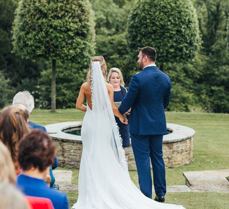 Bride in Made With Love Wedding Dress and Groom in Navy Suit Exchanging Vows at The Outdoor Wedding Ceremony