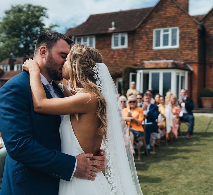 Bride in Backless Made With Love Wedding Dress and Lace Edge Cathedral Length Veil Kissing Her Groom in a Navy Suit at the Altar