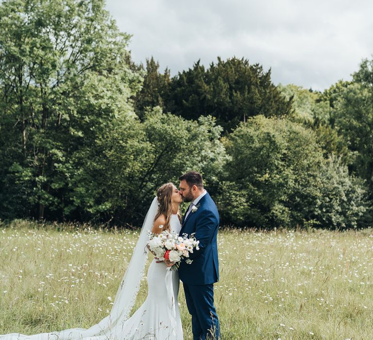 Bride in Halterneck Made With Love Wedding Dress and Groom in Navy Suit Kissing In a Field