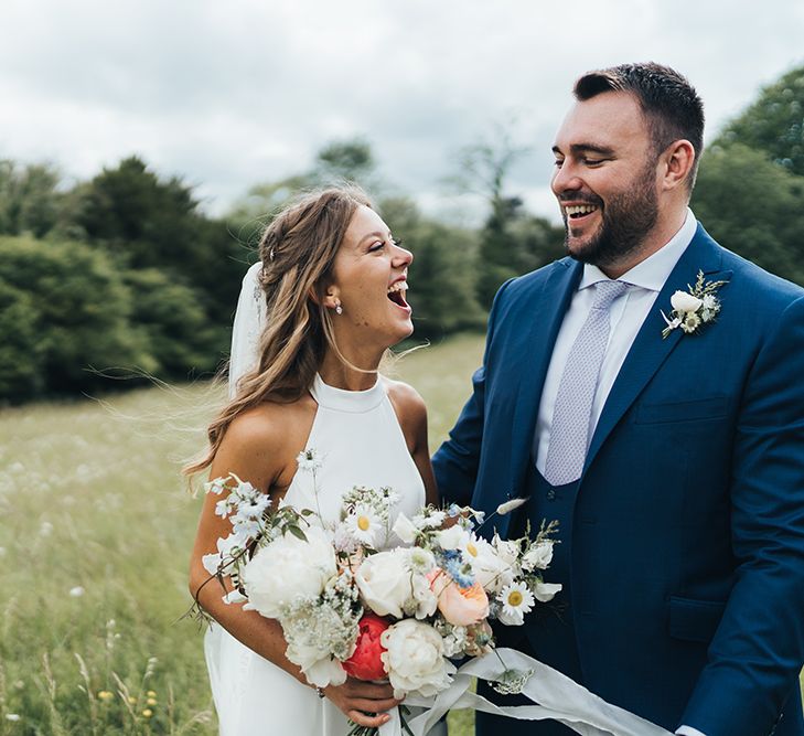 Bride in Halterneck Wedding Dress Holding a Pastel Wedding Bouquet Laughing with Her Groom in a Field