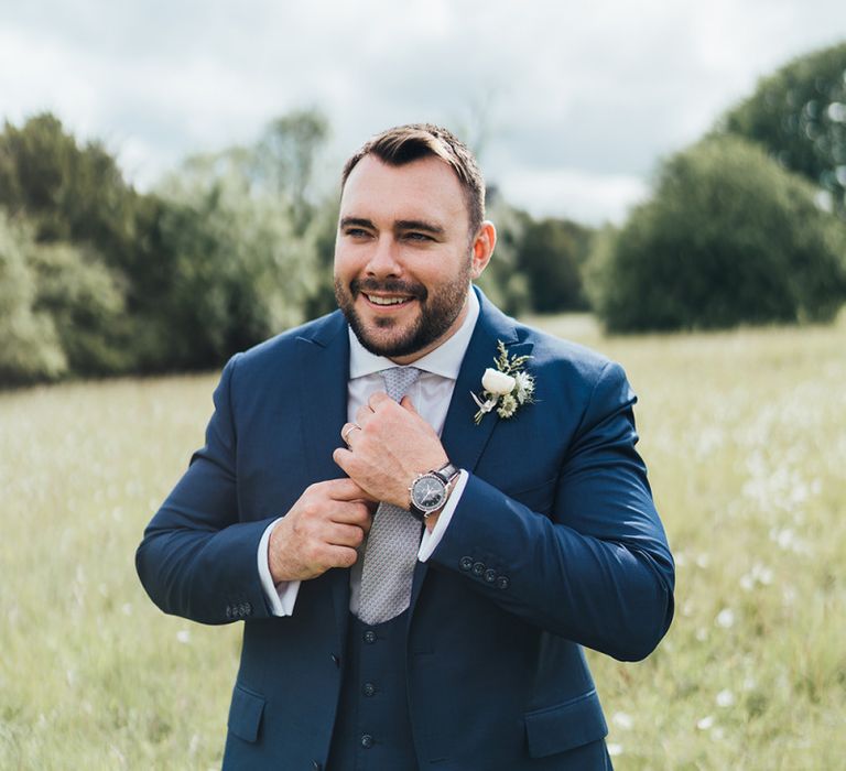 Groom in Three Piece Navy Suit with Horseshoe Waistcoat Adjusting His Tie in a Field