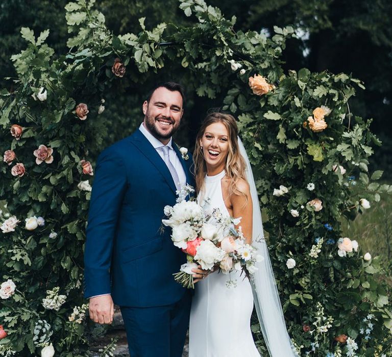 Bride in Fitted Wedding Dress Holding a Pastel Bouquet and Groom in Navy Suit Standing in Front of a Flower Arch