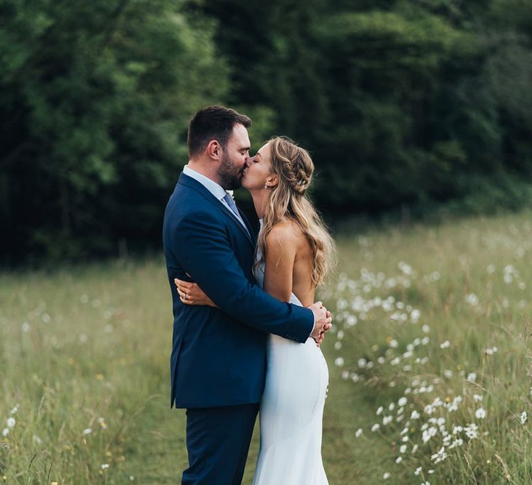 Bride in Halterneck Wedding Dress and Groom in Navy Suit Kissing in a Field