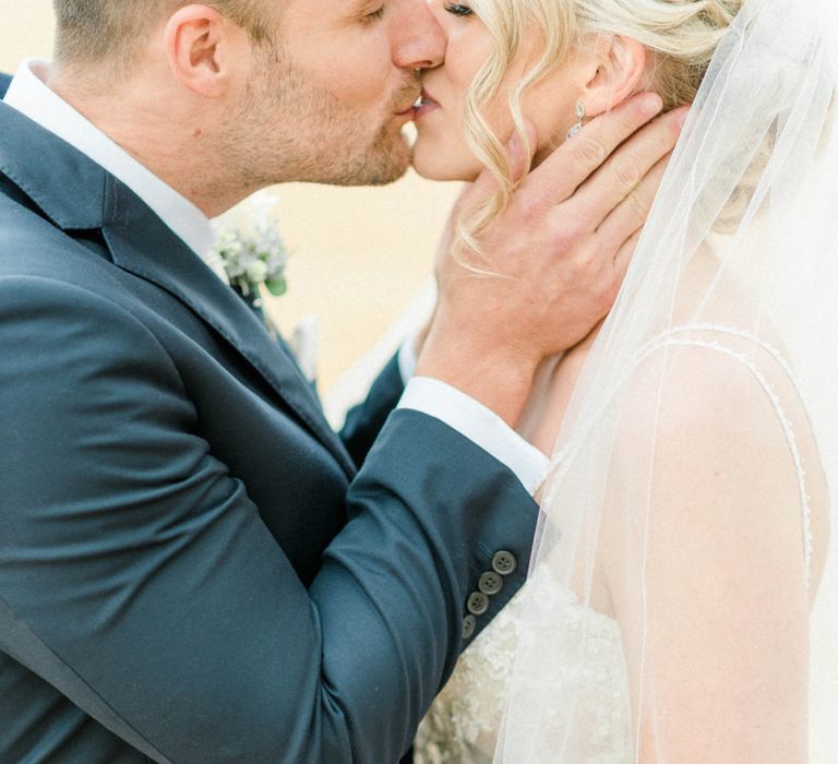 Groom in Moss Bros. Navy Suit Kissing his Bride in Lace Wedding Dress and Cathedral Veil