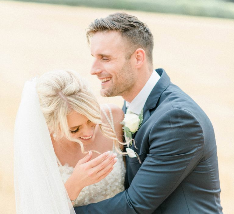 Bride in Lace Wedding Dress and Cathedral Veil laughing with Her Groom in  Navy Moss Bros. Suit