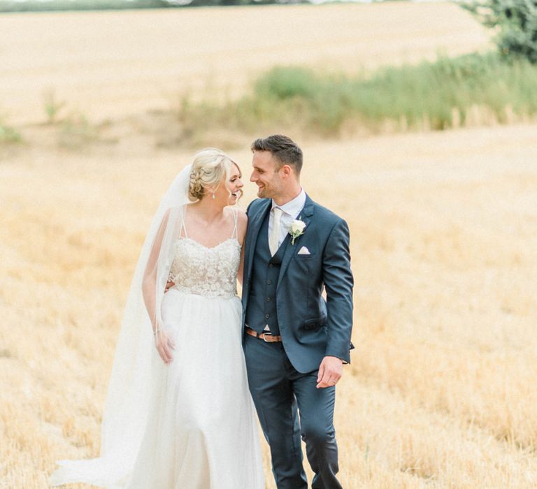 Bride in Lace Wedding Dress and Cathedral Veil and Groom in  Navy Moss Bros. Suit Walking Through a Corn Field