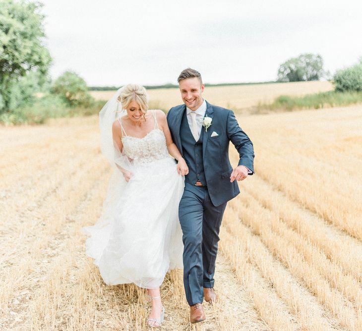 Bride in Lace Wedding Dress and Cathedral Veil and Groom in  Navy Moss Bros. Suit Holding Hands in a Corn Field