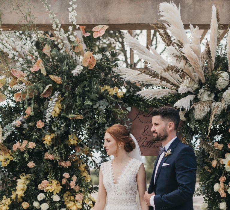 Bride in Pronovias Wedding Dress and Groom in Navy Suit Holding Hands in Front of a Peach Floral Arch