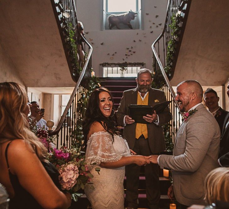 Bride in Off the Shoulder Grace Loves Lace Wedding Dress and Groom in Grey Blazer and Bow Tie Holding Hands at the Wedding Ceremony