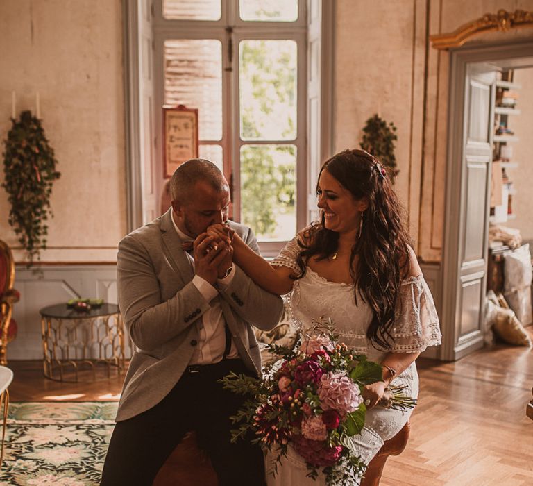 Groom in Grey Blazer and Bow Tie Kissing His Brides Hand in an Off The Shoulder Wedding Dress