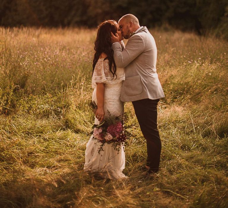 Bride in Emanuela Grace Loves Lace Wedding Dress and Groom in Grey Blazer Kissing in a Field