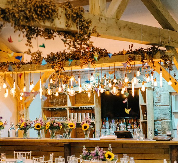 Eddison Light Hanging Bar Decoration | Paper Cranes Hanging from Barn Beams | Sunflower Bouquets in Vases | Colourful Paper Cranes &amp; Sunflower Wedding Décor in Rustic Barn | Sarah-Jane Ethan Photography