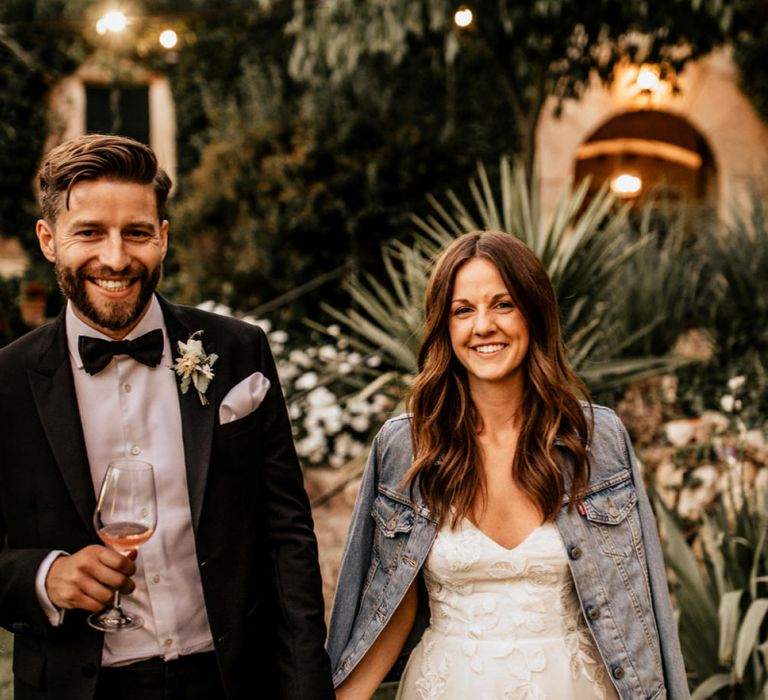 Bride in Tiered Lace Emma Beaumont Wedding Dress and Denim Jacket and Groom in Givenchy Tuxedo Laughing Whilst Holding Hands