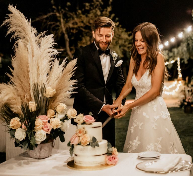 Bride in Tiered Lace Emma Beaumont Wedding Dress and Denim Jacket and Groom in Givenchy Tuxedo Cutting the Cake