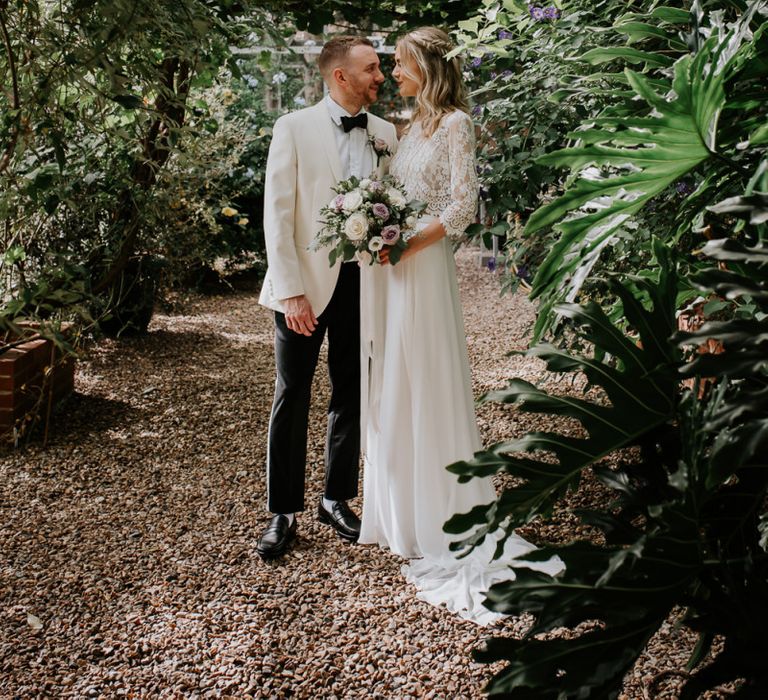 Bride in Wendy Makin Bridal Separates and Groom in White Dinner Jacket Embracing Under  Some Greenery