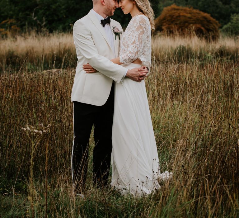 Bride in Lace Top and Plain Skirt by Wendy Makin  with Groom in White Dinner Jacket and Bow Tie Embracing in a Field