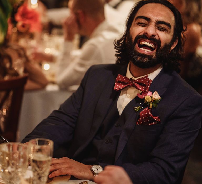 Groomsman in Navy Suit and Red Bow Tie Laughing During Wedding Reception Speeches