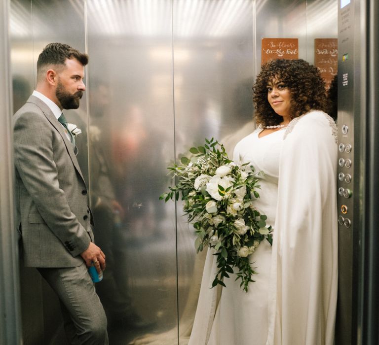 Portrait of bride in a floor length bridal cape and groom in grey suit inside a lift