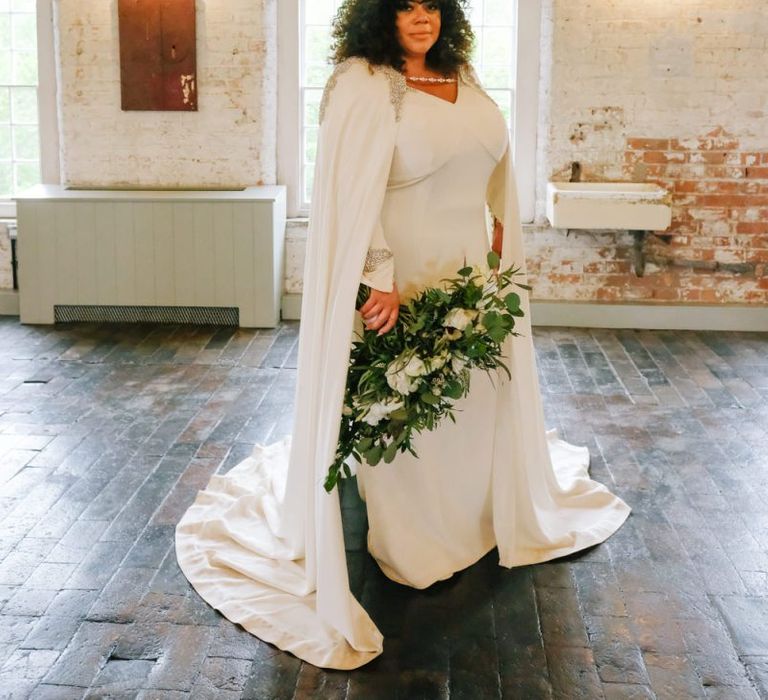 Bride in bridal cape over sleeved dress with curly hair down and white flower bouquet