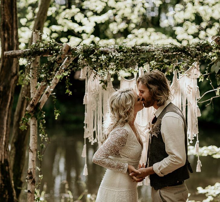 Bride in Lace Wedding Dress and Groom in Waistcoat Kissing at an Outdoor Woodland Wedding Ceremony