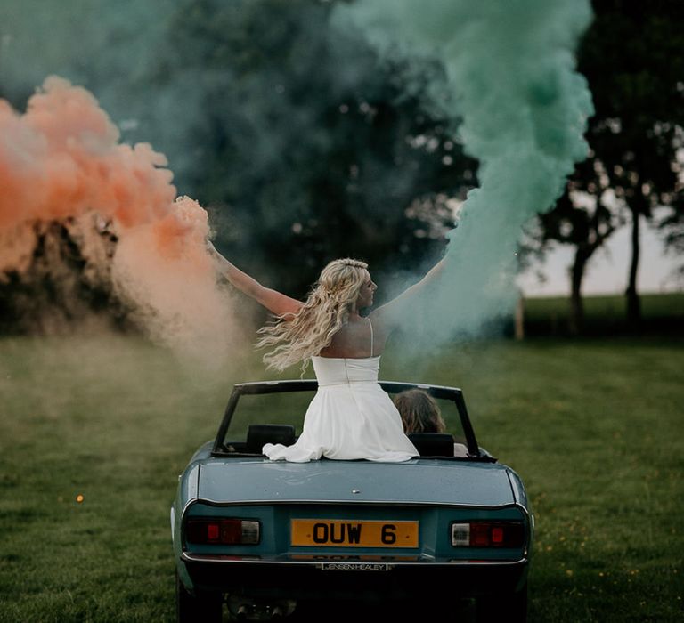 Bride Sitting in a Vintage Wedding Car Holding Coloured Smoke Flares